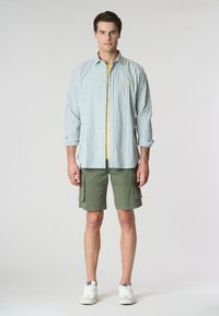 Camicia a righe con strisce verdi e bianche sopra una maglietta gialla, abbinata a pantaloni cargo verde oliva e sneakers bianche.