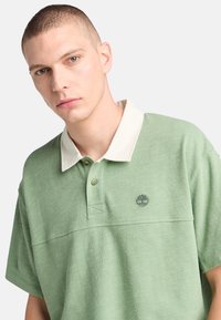 Grön bomullspolo med vit krage, två knappar och en trädlogotyp broderad på vänster bröst. Texturerad tyg med korta ärmar.