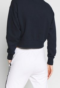 Felpa cropped blu navy con scollatura rotonda e maniche lunghe, caratterizzata da polsini e orlo a coste. Abbinata a pantaloni della tuta bianchi.