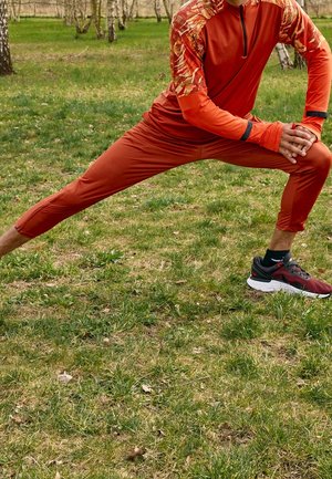 Czerwona sportowa bluzka z długim rękawem z wzorzystymi rękawami, w zestawie z dopasowanymi spodniami dresowymi i czerwono-czarnymi butami do biegania, na trawiastym terenie.