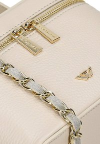 Gros plan d’un sac en cuir beige texturé avec des tirettes de fermeture éclair dorées et une bandoulière chaîne dorée entremêlée de suède beige.