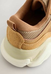 Brązowe sneakersy z siateczkową cholewką i zamszowymi akcentami. Okrągła, masywna podeszwa w kremowym kolorze, z pętelką i kontrastującymi przeszyciami.
