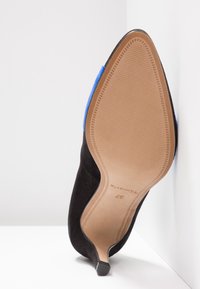 Chaussure à talon haut pour femme noire à bout pointu, avec semelle beige et accent bleu, présentée debout avec la semelle face au spectateur.