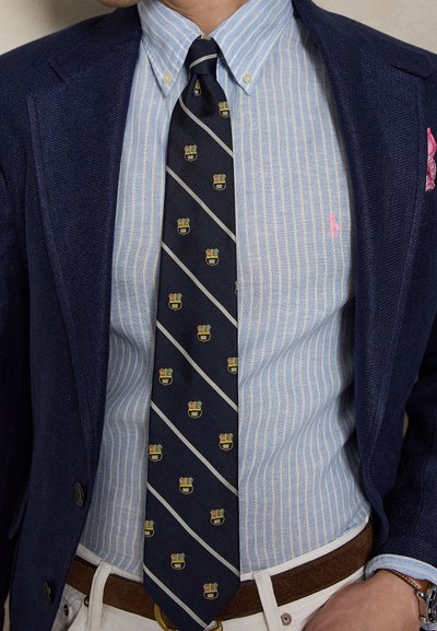 Mees kannab tumesinist bleiserit, hele sinist triibulist nööpidega särki, tumesinist lipsu vapimustritega, valgeid pükse ja pruuni vööd.