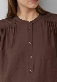 Bruine, lichte blouse met een ronde halslijn, knoopsluiting aan de voorkant en geraffineerde details bij de schouders. Gladde textuur, subtiele glans.