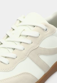 Λευκό αθλητικό παπούτσι με μπεζ σουέντ λεπτομέρειες, διαθέτοντας υφή στην επάνω επιφάνεια, στρογγυλεμένη μύτη και λευκά κορδόνια. Η σόλα από καουτσούκ προσφέρει πρόσφυση και ανθεκτικότητα.