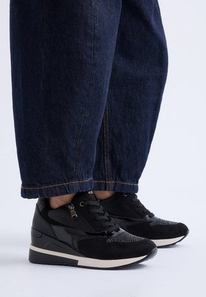 Pies con zapatillas negras con suelas blancas y vaqueros anchos de pierna azul oscuro sobre un fondo claro y liso.