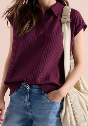 Woman wearing a ribbed dark purple short-sleeve polo shirt with blue jeans, holding a light beige shoulder bag strap, hand in pocket.