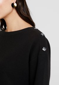 Svart texturerad tröja med rund halsringning och tre stora svarta knappar på axeln. Kompletterad med runda svarta och guld örhängen.