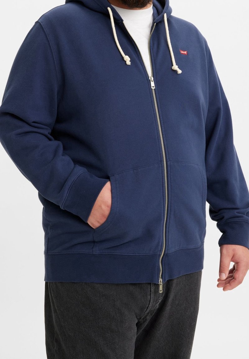 Homme portant un sweat à capuche zippé bleu marine avec des cordons écrus et les mains dans les poches, assorti à un pantalon noir sur un fond uni.