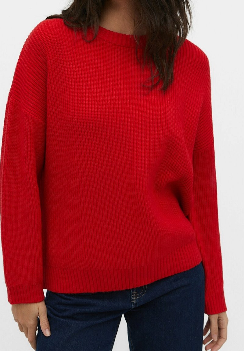Femme portant un pull en maille côtelée d'un rouge vif à manches longues et un jean bleu foncé, debout devant un fond blanc.
