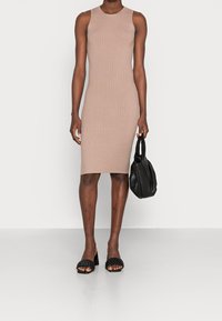 Robe beige sans manches en maille à texture côtelée, longueur au genou. Le modèle tient un sac à main noir doux et porte des sandales tissées noires.