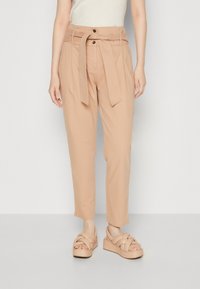 Helle Pfirsichfarbene Hose mit hoher Taille, Stoffgürtel, zwei aufgesetzten Taschen und schmal zulaufendem Bein. Kombiniert mit klobigen beigen Plateau-Sandalen.
