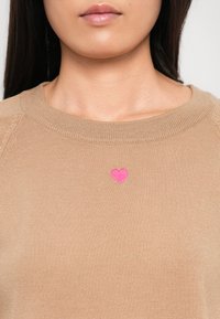 Kvinne iført beige genser med liten rosa hjertebrodering sentrert under utringningen, mot en ensfarget lys bakgrunn.