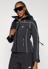 Casaco softshell preto com costuras refletivas, bolsos com fecho de correr, capuz e punhos elásticos. Usado com calças escuras e grandes óculos de sol pretos.