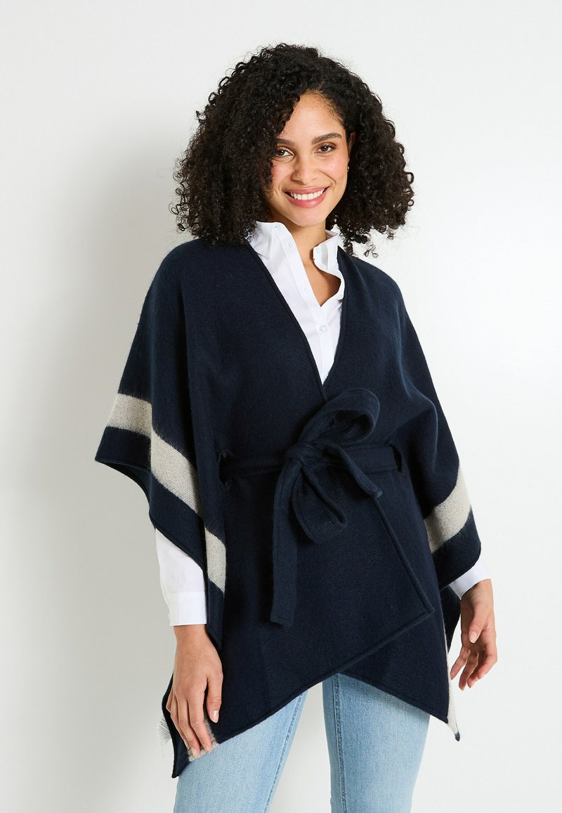 Poncho azul marino hecho de una tela suave con un cinturón de lazo. Presenta rayas diagonales en color crema y un escote en V. Lleva puesto sobre una camisa blanca y vaqueros.