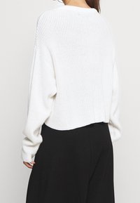 Pull blanc court en tricot avec une texture côtelée, des manches larges et un ourlet arrondi. Caractérisé par une coupe décontractée et des détails de couture subtils.