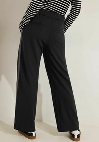 Zwarte wijde broek met een zachte textuur, voorzien van een hoge taille en twee achterzakken, gedragen met witte sneakers.