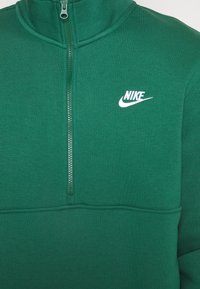 Tmavě zelená mikina Nike s polovičním zipem a hladkou texturou, s bílým logem Nike v horním levém rohu.