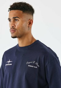 Jonge man met een curly fade kapsel, gekleed in een marineblauwe shirt met de tekst "Project X Paris Members' Club" en een kruisoorbel, op een witte achtergrond.