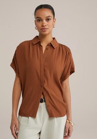 Camicia a maniche corte marrone con un motivo testurizzato. Presenta un colletto e una vestibilità ampia, abbinata a pantaloni chiari e una cintura nera.