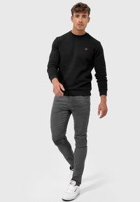 Schwarzer Sweatshirt mit Struktur, rundem Ausschnitt und braunem Logo-Patch, kombiniert mit grauen Slim-Fit-Jeans mit gesteppten Knieakzenten.