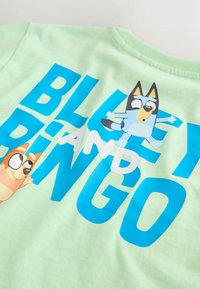 Mintgrøn skjorte med stort blåt tekst "BLUEY AND BINGO" og tegneseriehundfigurer, en der hopper og en der kigger frem fra siden.