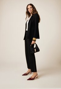 Zwarte op maat gemaakte blazer over een witte blouse, zwarte broek, gecombineerd met een zwarte handtas en bordeauxrode lakleren hakken. Eenvoudige gouden armbanden om de pols.