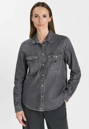 Vrouw met lang bruin haar, die een verweerde zwarte spijkerblouse met knopen en een donkere broek draagt, staand tegen een effen lichte achtergrond.