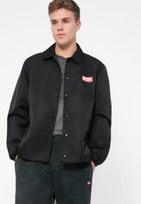 Junger Mann trägt eine schwarze knöpfbare Jacke mit rotem Diesel-Logo auf der Brust und dunkelgraue Jogginghose mit passendem Logo am Bein.