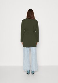 Olive green long coat with a belt, worn over light blue wide-leg jeans, paired with teal heels. Smooth fabric, minimal detailing.
