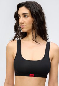 Γυναίκα με μακριά, κατσαρά μαλλιά που φοράει μαύρο αθλητικό σουτιέν με κόκκινο λογότυπο Hugo, κοιτάζοντας προς τα πλάγια σε φόντο απλό.