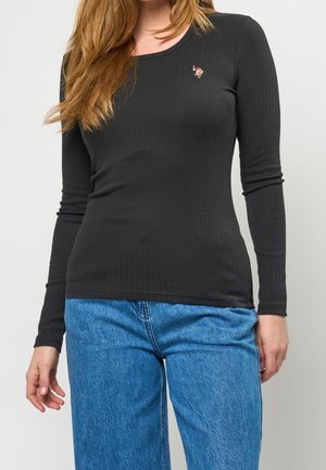 Kvinde iført en tætsiddende sort ribstrikket langærmet skjorte med et lille broderet logo, kombineret med løse blå cowboybukser.