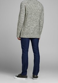 Gestrickter grüner Pullover mit mehrfarbigen Sprenkeln, dunkelblaue Jeans und graue Turnschuhe mit weißen Akzenten, von hinten gesehen.