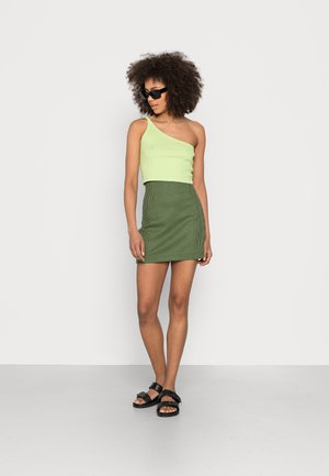 Top cropped verde limão de um ombro combinado com uma saia mini xadrez verde. Sandálias pretas completam o visual. Texturas suaves e design ajustado.