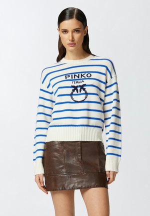 Maglione color crema con righe orizzontali blu, presenta un logo sul petto, abbinato a una mini gonna in pelle marrone con toppe testurizzate.