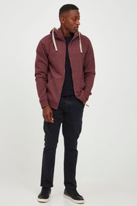 Kardinalrød hættetrøje med lynlås og grå snore, sidelommer, båret over en sort top, kombineret med mørke bukser og sorte sneakers.