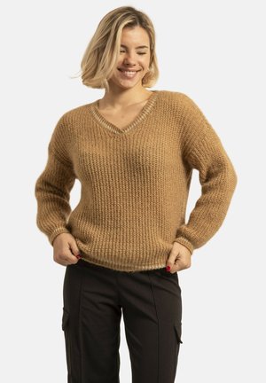 Femme souriante aux cheveux blonds courts porte un pull en tricot marron et un pantalon noir, tenant l'ourlet du pull avec les deux mains.