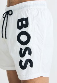 Bijele kupaće gaće s crnim "BOSS" logotipom sa strane, sa elastičnom trakom u struku i vezicom za podešavanje.
