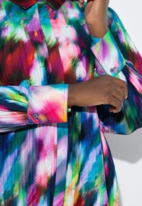 Farverig plisseret kjole med krave; har levende mønstre i pink, blå, grøn og lilla. Knapdetalje på manchetterne.