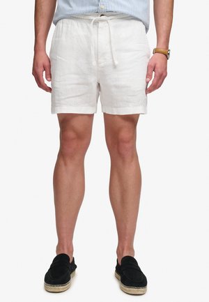 Mies, joka käyttää valkoisia nyörillisiä shortseja, mustia slip-on kenkiä, sinisävyistä raitapaitaa housujen sisällä ja ruskeaa kelloa vasemmassa ranteessa.