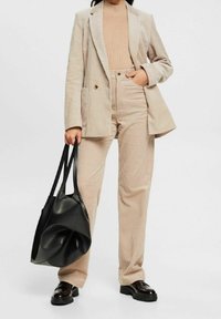 Person wearing beige corduroy blazer and pants, tan ribbed top, black loafers, holding large black leather tote bag by side.
