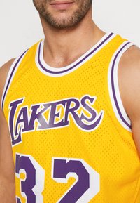 Maglia da basket gialla con il testo "LAKERS" in viola e dettagli bianchi. Presenta una texture a rete e il numero "32" in evidenza.