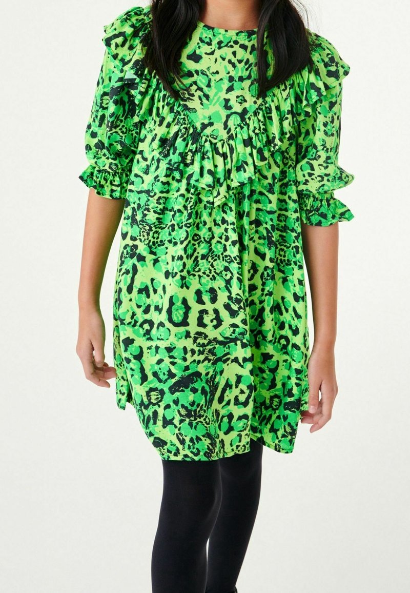 Fille portant une robe à imprimé animal vert fluo et noir avec des volants sur la poitrine et les manches, associée à des leggings noirs.