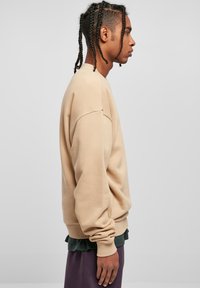 Felpa beige con vestibilità rilassata, polsini e orlo a costine. Il modello indossa un sottogiacca verde scuro e pantaloni viola scuro.