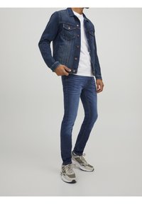 Chaqueta de mezclilla en azul oscurecido con bolsillos frontales, combinada con jeans azules ajustados. Llevada con zapatillas gris y blancas, con detalles texturizados.