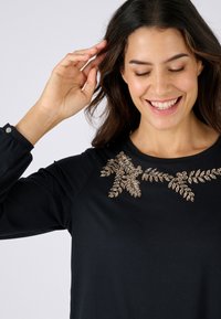 Maglietta nera a maniche lunghe con decorazione floreale di perline e paillettes sul petto, collo rotondo e dettaglio con bottone sul polsino.