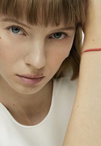 Primo piano di una persona con pelle chiara, che indossa un top bianco. Un bracciale rosso con design intrecciato è visibile sul suo polso.