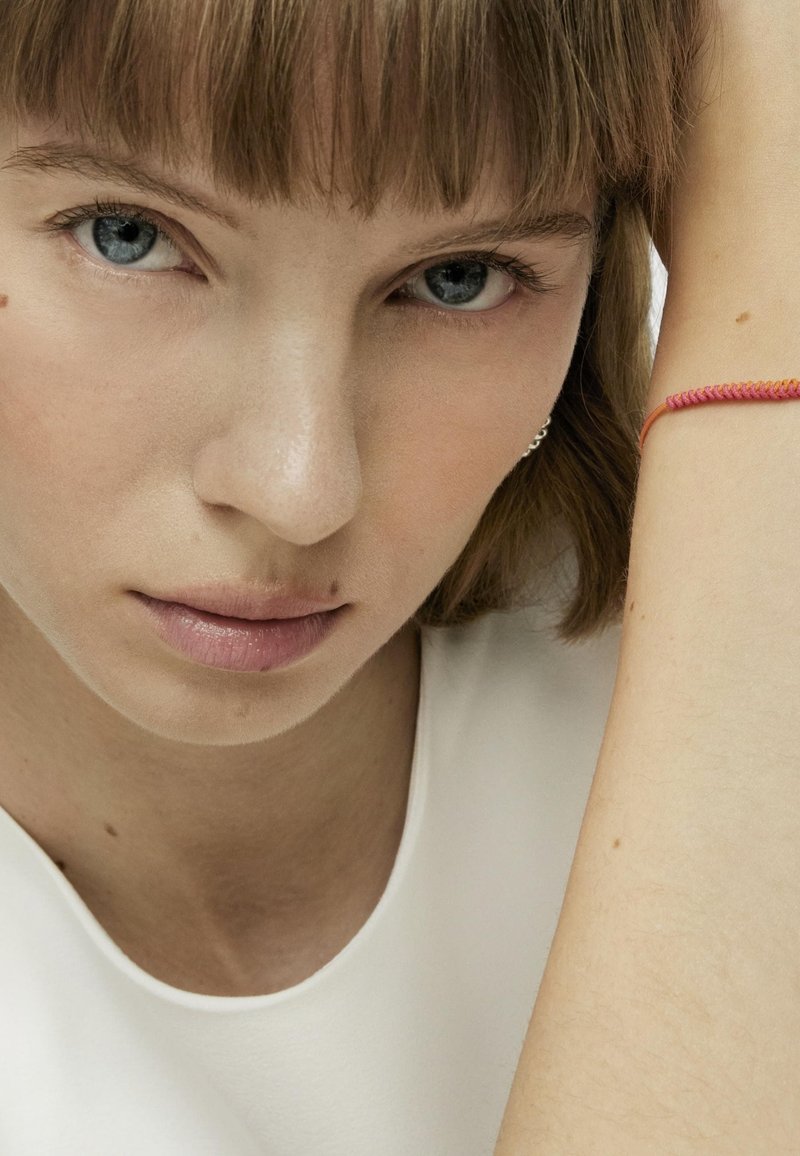 Primo piano di una persona con pelle chiara, che indossa un top bianco. Un bracciale rosso con design intrecciato è visibile sul suo polso.