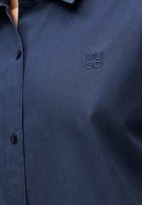 Κοντινό πλάνο ενός ατόμου που φοράει πουκάμισο σε σκούρο μπλε με κουμπιά, με το λογότυπο "HUGO" κεντημένο στο στήθος.